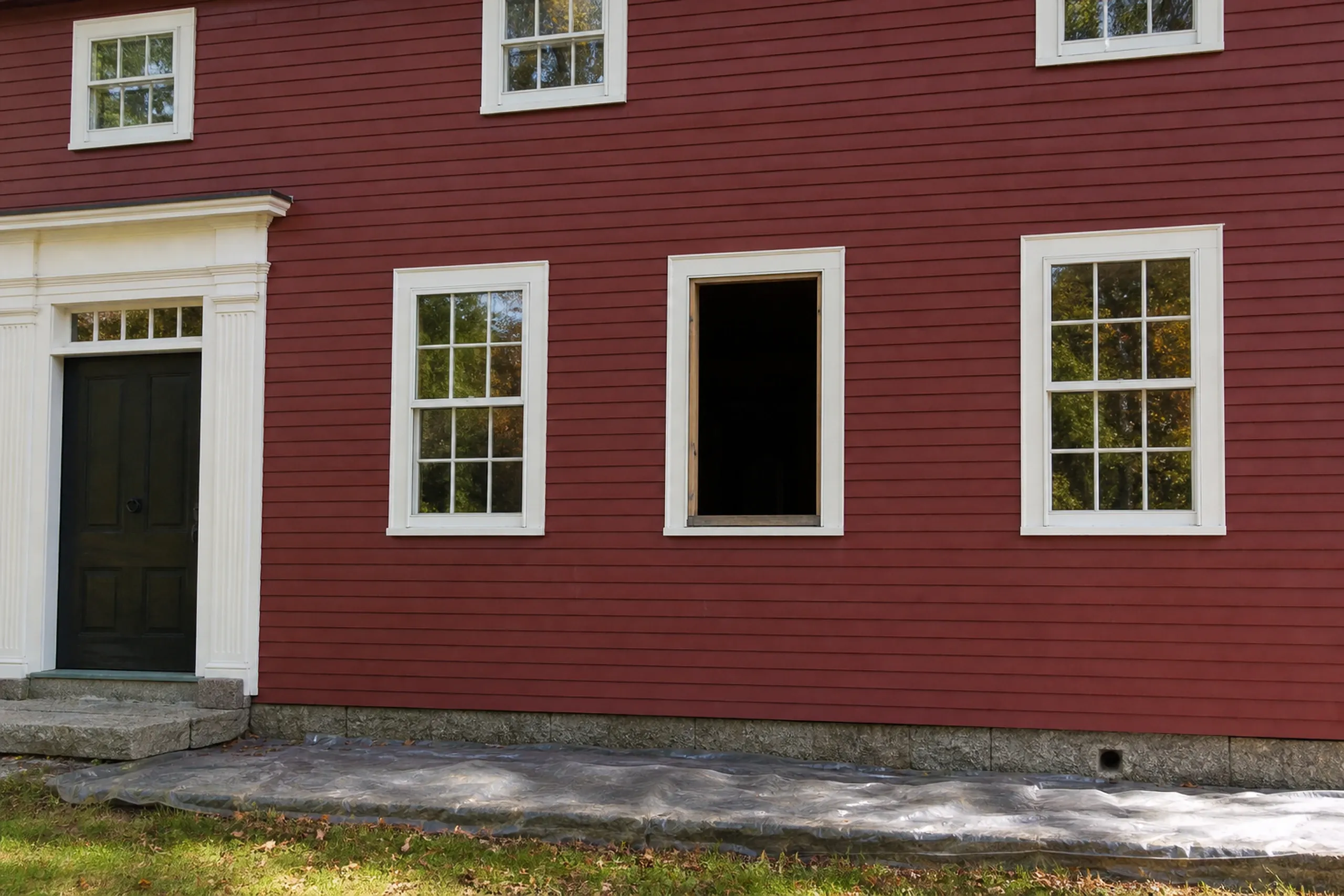 Window replacement in Friendship in progress: empty window opening in a bare wood frame between two white-trimmed windows on a red clapboard wall, plastic sheeting on the grass