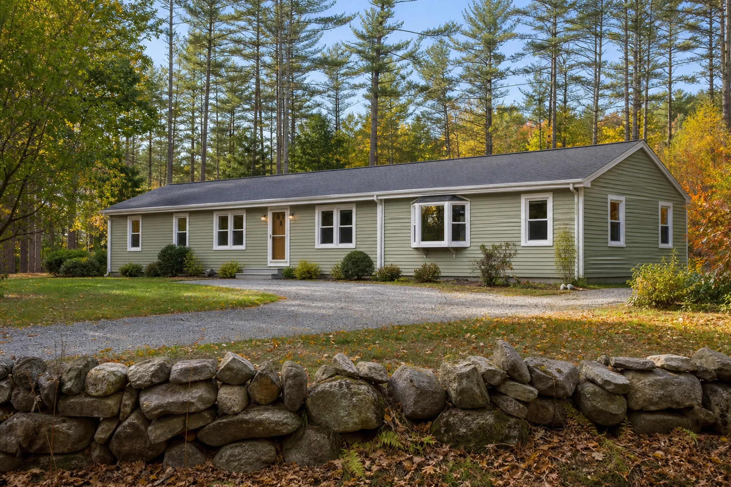 Window replacement in Searsmont: long one-story house with sage green siding, white-trimmed windows and a bay window behind a gravel drive and stone wall, under tall pines